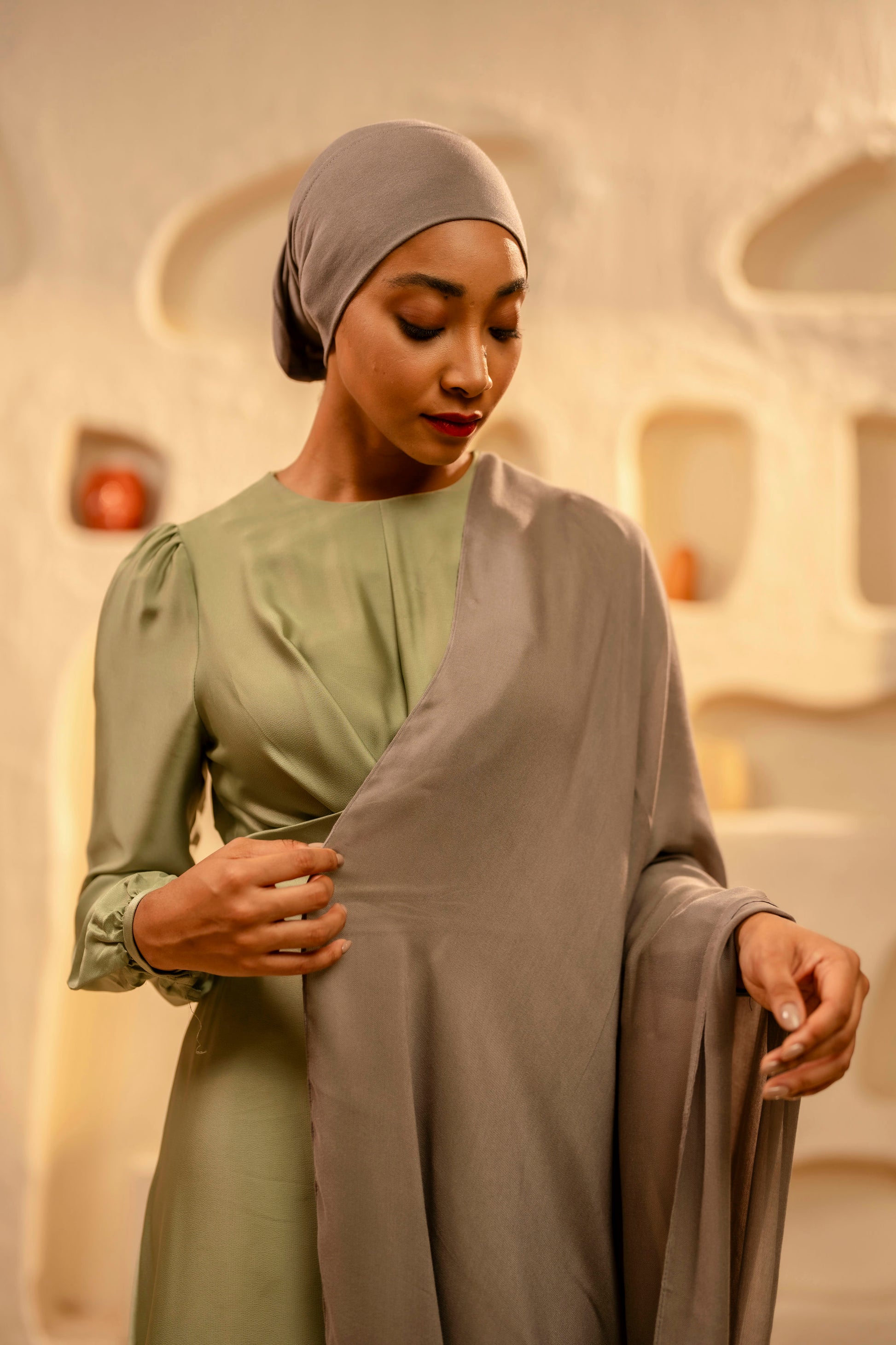 Woman wearing a green dress and khaki headscarf in an indoor setting with decorative elements.