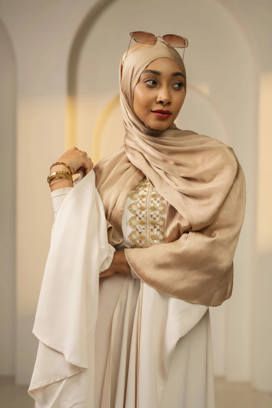 Woman wearing a gold hijab and white dress with gold patterns indoors.