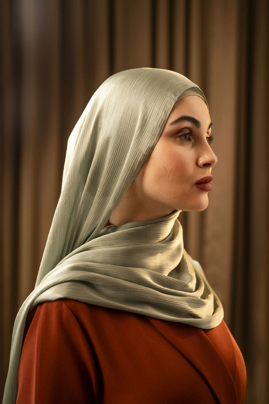 Woman wearing a light green silk hijab and red garment against a brown curtain background