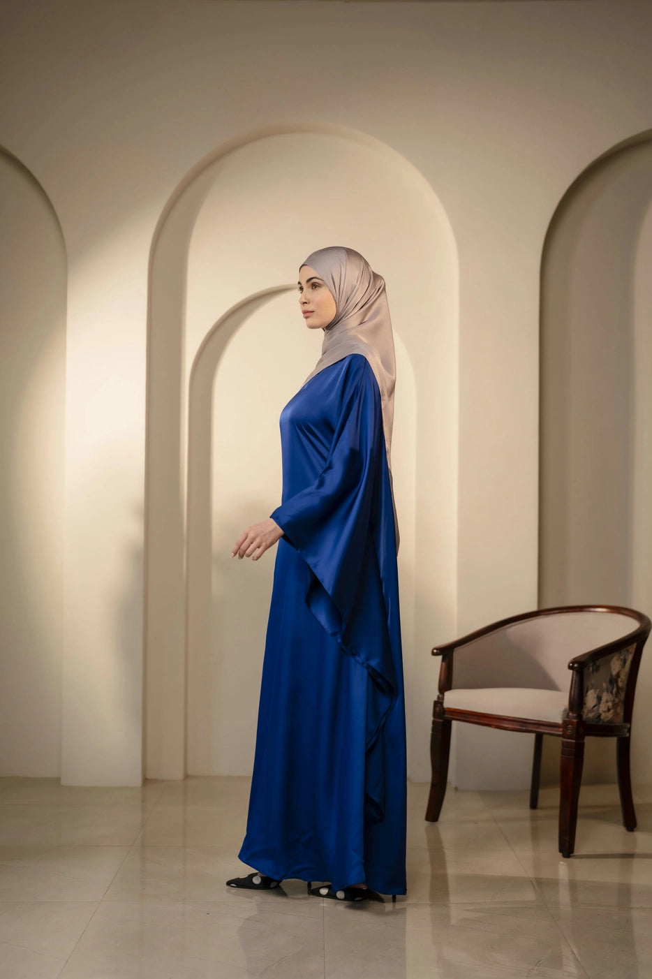 Woman wearing silver gray headscarf in a navy dress standing in a room with arches and a chair.
