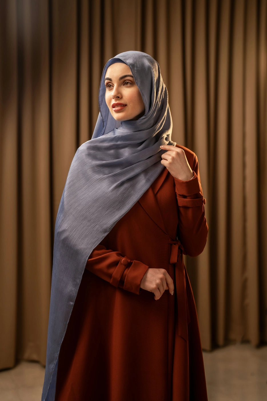 Woman wearing a shiny blue hijab and red dress against a brown curtain background