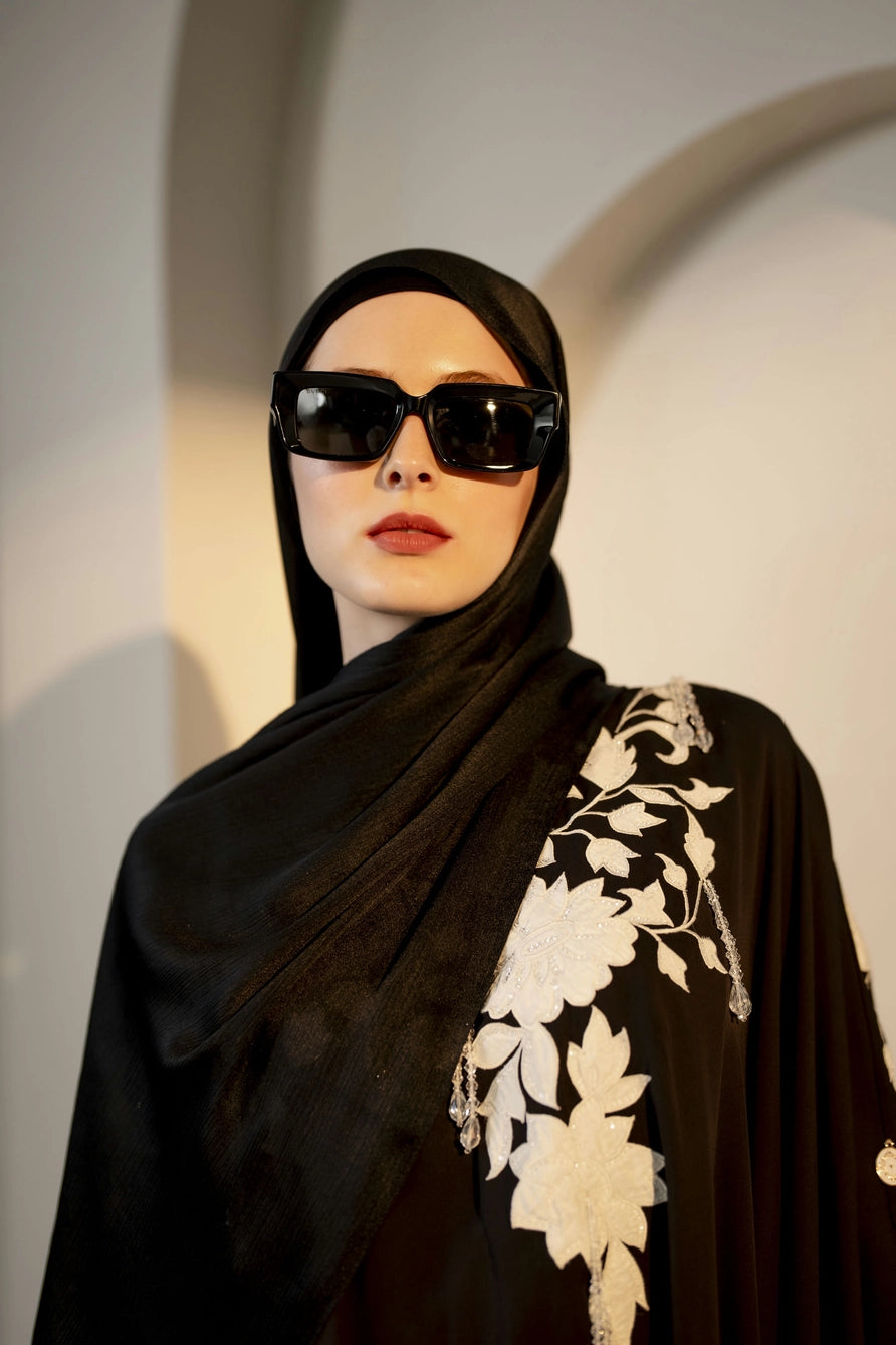 Person wearing a black abaya with white floral patterns and sunglasses against a neutral background ready to celebrate eid
